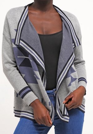 Personne portant un cardigan à motifs gris et bleu ouvert devant, un top noir et un jean bleu, ajustant le cardigan à la taille.