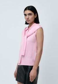 Lys rosa ermeløs strikket topp med polo-krage, med et matchende strikket slips hengende over skulderen. Teksturert overflate.