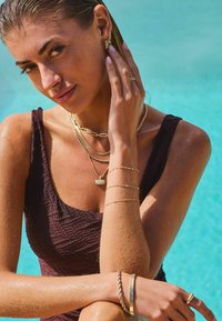 Les bijoux en or comprennent des colliers superposés, des boucles d'oreilles créoles et des bracelets. Le mannequin porte un maillot de bain texturé, marron foncé, au bord d'une piscine.