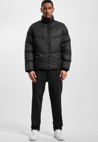 Sort pufferjakke med høj krave, lynlås foran og quiltet design. Båret over sorte bukser og hvide sneakers. Enkle detaljer hele vejen igennem.
