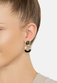 Gold and black earrings featuring a green stone, with an oval hoop design and textured gold accents. Glossy and smooth finish.
