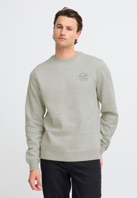 Sudadera gris claro con cuello y puños acanalados, que presenta un pequeño logotipo impreso en el pecho. Combinada con jeans oscuros.
