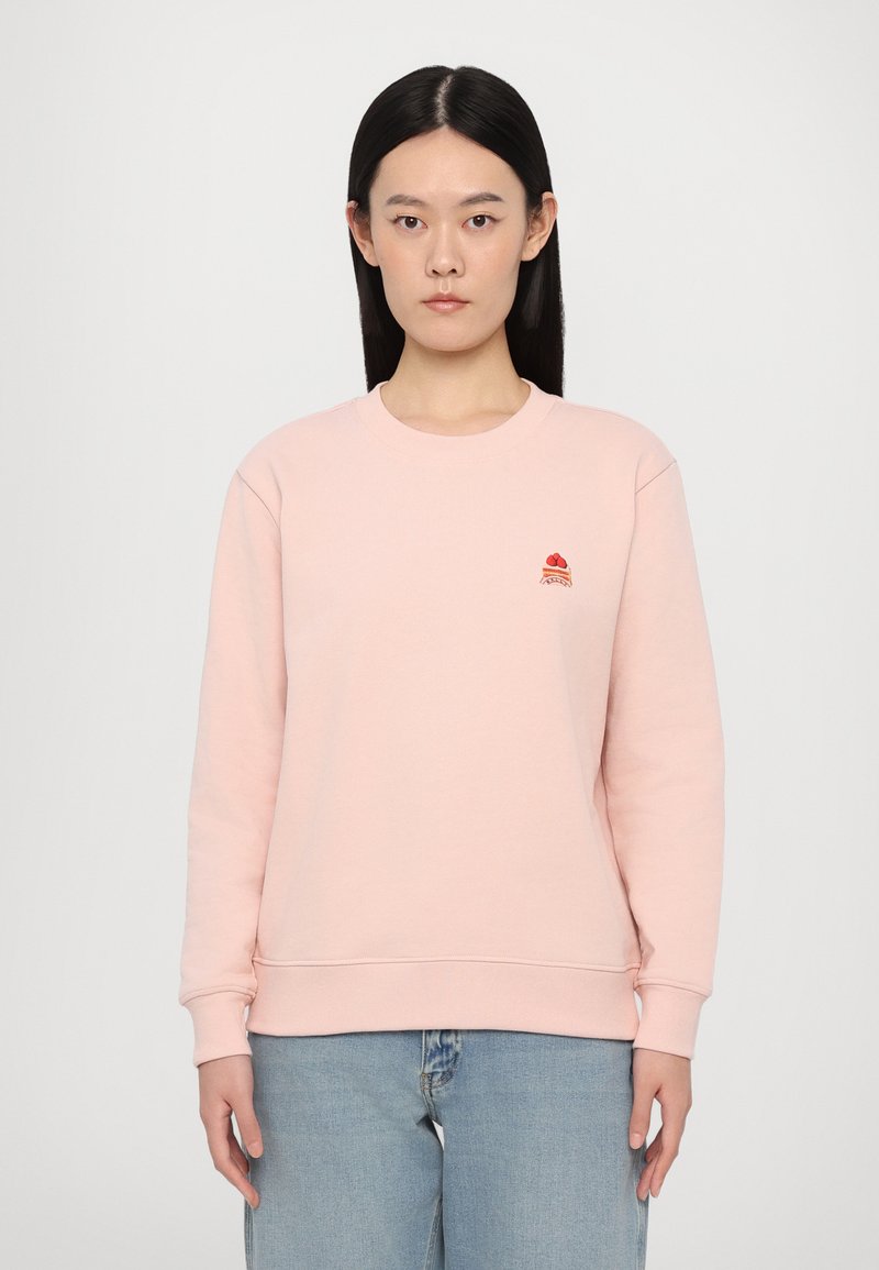 Kvinde med langt sort hår iført en lyserød sweatshirt med lille rød broderet design på brystet og lyseblå jeans, neutral baggrund.