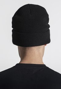 Berretto di lana nera lavorato a maglia con texture a coste, vestibilità aderente e visiera pieghevole, visto da dietro. Indossato da una persona con una maglietta nera.