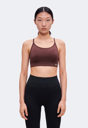 Femme aux cheveux raides et foncés portant un soutien-gorge de sport marron et des leggings noirs taille haute, debout devant un fond clair uni.