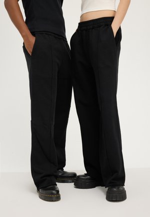 Deux personnes portant des pantalons noirs à jambes larges et des chaussures noires épaisses, debout côte à côte avec les mains dans les poches.