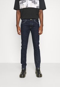 Granatowe jeansy o wąskim kroju, z przednimi kieszeniami, zapięciem na guziki oraz subtelnymi detalami przeszyć. Noszone z czarnymi butami.