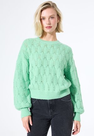Mujer rubia con un suéter de punto texturizado en verde menta con mangas de globo y jeans negros con manchas, frente a un fondo blanco.