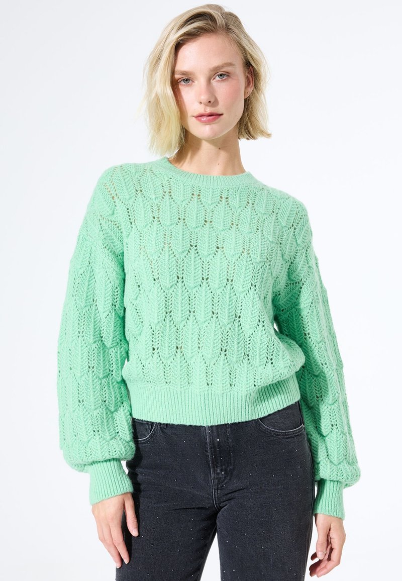Blonde vrouw in een mintgroen textuur gebreid trui met ballonmouwen en zwart met witte gespikkelde jeans tegen een witte achtergrond.