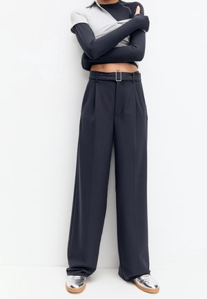 Persoon draagt zwarte wijde broek met hoge taille, zilveren schoenen, zwart topje met lange mouwen onder korte grijze mouw, armen gekruist.