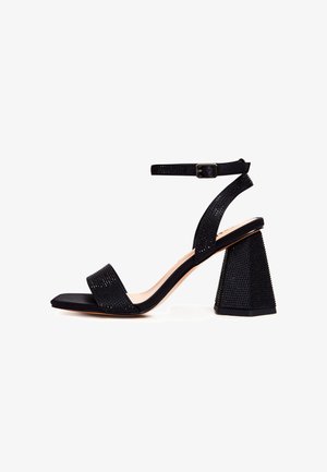 Sort sandal med åben tå, ankelrem, blokhæl og små nittebesætninger på remme og hæl.