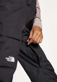 Juodi nepralaidūs vandeniui kelnių su šoninėmis užtrauktukais ir krovininėmis kišenėmis; turi "The North Face" logotipą baltos spalvos kairėje kojyje.