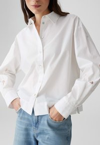 Witte button-up shirt van lichte stof, met subtiele uitsnijdingen op de mouwen en een klassieke kraag. Gecombineerd met lichtblauwe jeans.