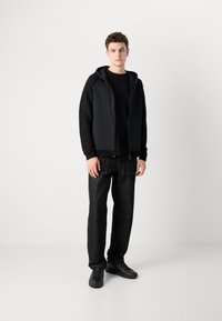 Črna kapuca z zadrgo z gladko površino, oblečena čez črno majico. Kombinirana z ravno krojenimi temnimi kavbojkami in črnimi teniskami.