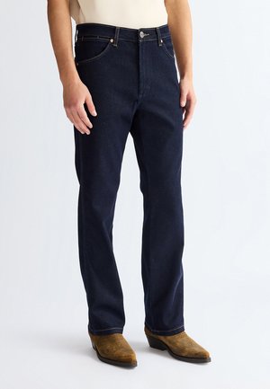 Mies, jolla on päällään tummansiniset suoralahkeiset farkut ja ruskeat cowboy-saappaat, seisoo valkoista taustaa vasten.