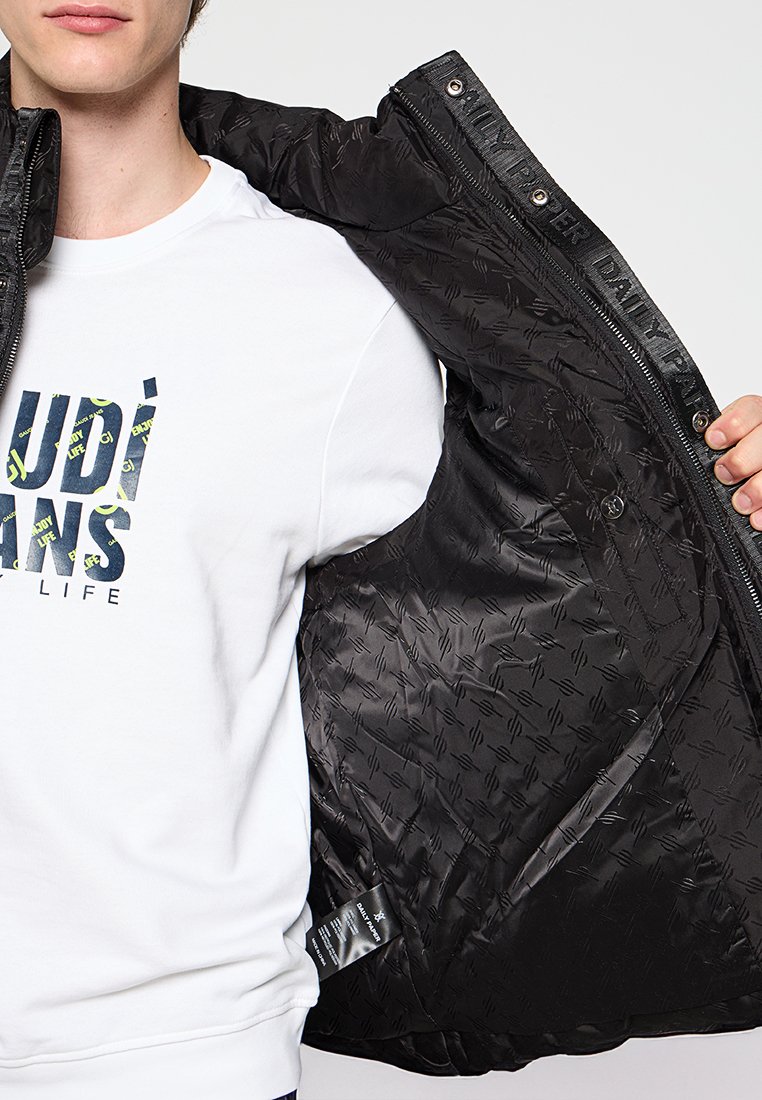 Zwarte gewatteerde jas met een diamantquiltpatroon en binnenvoering. Heeft een rits en een label aan de binnenkant, gedragen over een wit shirt.