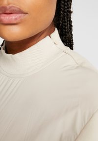 Beige Hochkragenoberteil mit gerippter Textur, das einen zulaufenden Kragen und subtile Knopfdetails aufweist. Glatter Stoff mit weichem Fall.