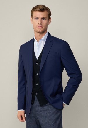 Blazer bleu marine à coupe ajustée, associé à un gilet noir avec des accents de boutons, et une chemise à carreaux bleu clair en dessous. Pantalons texturés gris.