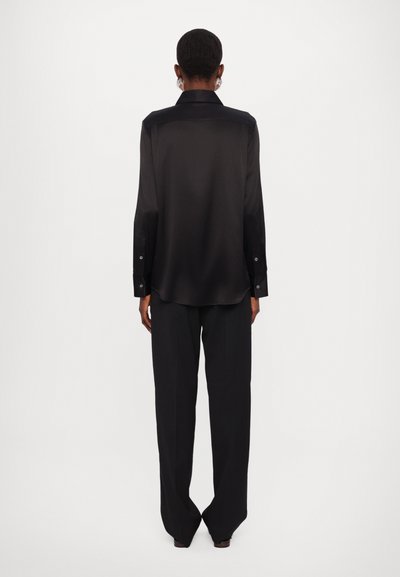 Chemise noire en satin à manches longues avec un col, des poignets boutonnés et un ourlet légèrement incurvé. Accompagnée de pantalons noirs ajustés.