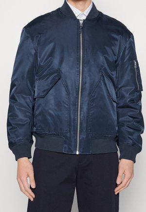 Persona que lleva una chaqueta bomber azul marino con cremallera frontal, bolsillos con solapa y puños acanalados, combinada con pantalones negros y una camisa blanca con cuello.