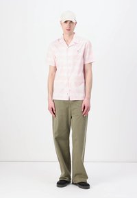 Camicia rosa a maniche corte con motivo geometrico, abbinata a pantaloni larghi color verde oliva e scarpe nere, indossata con un cappellino chiaro.