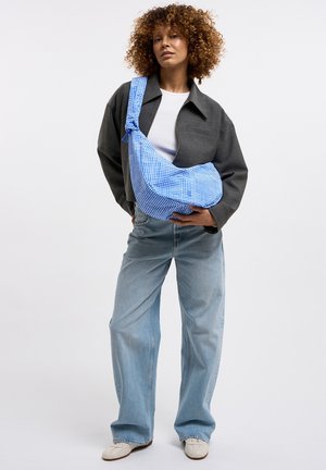 Mujer con cabello rizado que lleva una chaqueta gris, camisa blanca, jeans anchos de pierna en azul claro, zapatos blancos y sostiene un bolso de hombro azul a cuadros.
