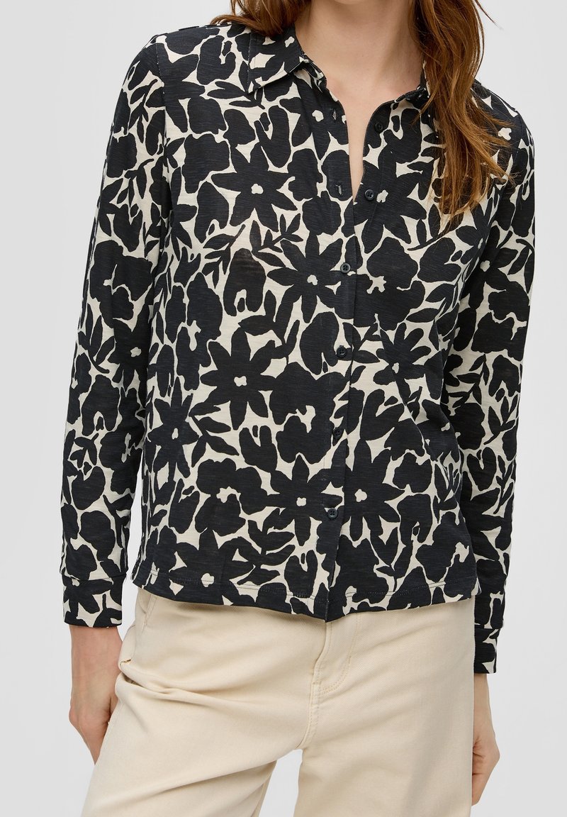 Femme portant une chemise à manches longues boutonnée à motif floral noir et blanc avec un pantalon beige, debout les mains relâchées le long du corps.