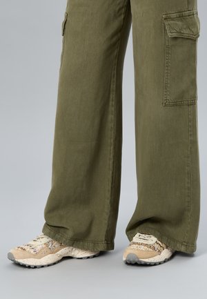 Beine, die olivgrüne, weite Cargohosen und beige gemusterte Sneakers mit strukturierten Sohlen auf grauem Boden tragen.