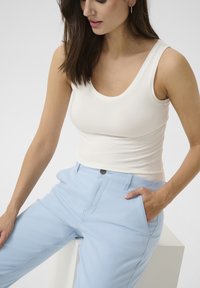 Bijela uska majica bez rukava u kombinaciji s svijetloplavim hlačama koje imaju bočnu džep i kopčanje na gumu u struku. Glatka tkanina, ležeran stil.