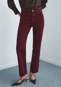 Personne portant un pantalon droit taille haute bordeaux et des escarpins à bout pointu noir, se tenant sur un tapis gris contre un mur blanc.