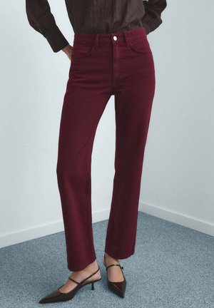 Persoon in hooggewaad bordeauxrode rechtpijpen broek en donkere puntige slingback hakken, staand op grijs tapijt tegen een witte muur.