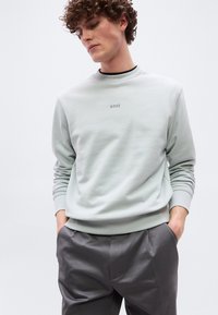 Νεαρός με σγουρά μαλλιά που φοράει ανοιχτό γκρι φούτερ με το κείμενο "BOSS" και σκούρο γκρι παντελόνι, με τα χέρια στις τσέπες, κοιτάζοντας προς τα κάτω.