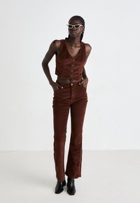 Woman wearing brown corduroy buttoned vest and matching pants with black boots, standing against a plain white background.