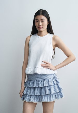 Ung kvinde med langt mørkt hår iført en hvid ærmeløs top og blå mininederdel med lagdelte flæser foran en ensartet grå baggrund.