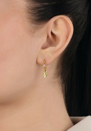 Goldene Creolen mit einem herzförmigen Anhänger, der ein strukturiertes Strahlenmuster aufweist und elegant vom Ohrläppchen hängt.