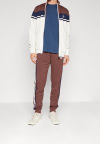 Herratletisk outfit bestående av en vit zip-up jacka med vinröda och marinblå ränder, i kombination med bruna byxor och ljusa sneakers.