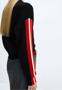 Musta neule, jossa on punaiset ja valkoiset raidoitukset hihoissa. Ominaisuus on istuva malli, ja se on yhdistetty harmaisiin housuihin.