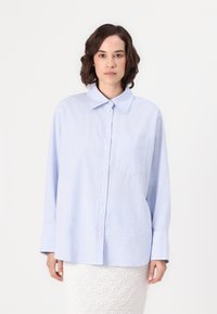Camisa de botão às riscas azuis e brancas, com colarinho, mangas longas e um único bolso no peito, apresentando um corte descontraído e uma textura macia.