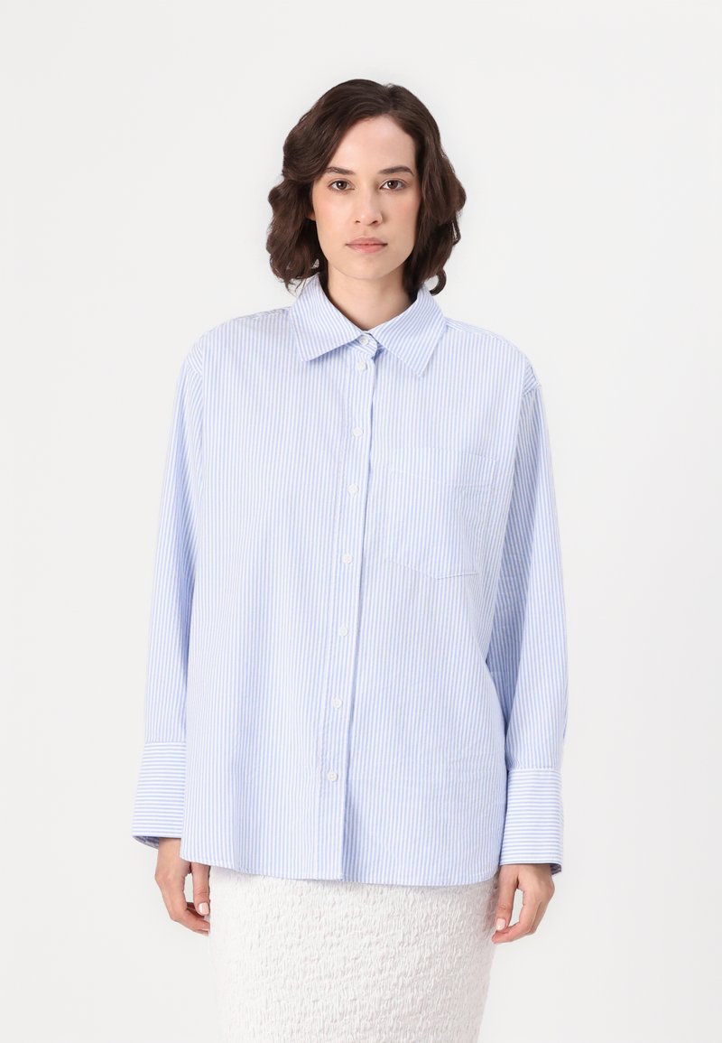 Camisa de botão às riscas azuis e brancas, com colarinho, mangas longas e um único bolso no peito, apresentando um corte descontraído e uma textura macia.