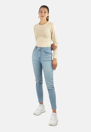 Beiges Langarmoberteil, hoch taillierte hellblaue Jeans und weiße Sneakers. Das Model steht mit Händen in den Hüften vor einem schlichten Hintergrund.