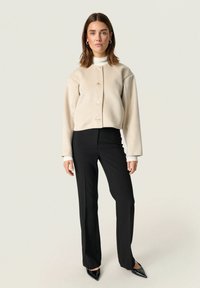 Beige cropped jacket with buttons, ribbed cuffs, and black fitted pants. Black pointed-toe shoes. Smooth texture, minimalist design.