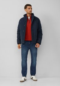 Marineblauwe pufferjack over een rode ritstrui, gecombineerd met blauwe jeans en witte sneakers. De jas heeft een hoge kraag en drukknoopsluiting.