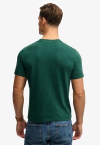 Grøn kortærmet T-shirt i bomuld med rund hals. Har en glat tekstur og en tætsiddende pasform, set bagfra. Kombineret med synlige jeans.