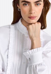 Blouse blanche avec un col haut à volants et un devant plissé. Les manches à poignets présentent des accents de boutons décoratifs. Texture de tissu lisse.