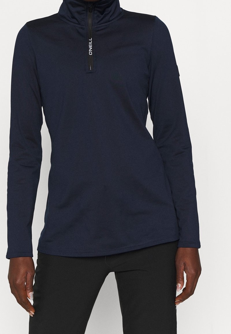 Persona che indossa una maglia a maniche lunghe blu navy con zip e il logo "O'Neill" sulla cerniera, abbinata a pantaloni neri, in piedi su uno sfondo semplice.