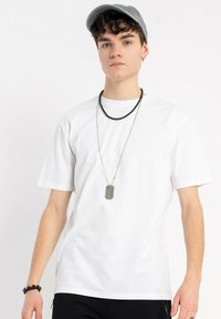 Biała bawełniana koszulka z krótkim rękawem, noszona z srebrnym naszyjnikiem z dog tag i czarnym naszyjnikiem z koralików, w zestawieniu z szarym czapką.