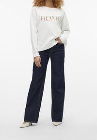 Vit sweatshirt med "FLOWER" broderat i blommönster. Kombinerad med mörkblå vida jeans och vita spetsiga skor.