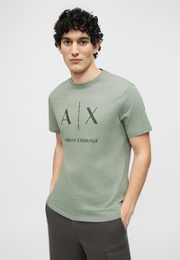 Νέος άνδρας με σγουρά μαλλιά, φοράει ένα ανοιχτό πράσινο T-shirt της Armani Exchange και σκούρα γκρι παντελόνια, με τα χέρια στις τσέπες, στέκεται μπροστά σε απλό φόντο.