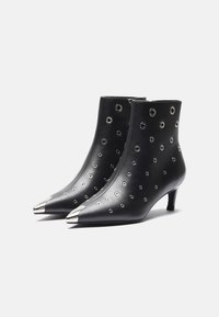 Botas de tobillo de cuero negro con puntas afiladas, tapas de acento plateadas y recortes circulares en la superficie, montadas sobre un tacón bajo.