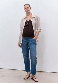 Mujer embarazada de pie contra una pared blanca, vistiendo una camisa a rayas, top negro, jeans azules, sandalias y un collar con un colgante circular.